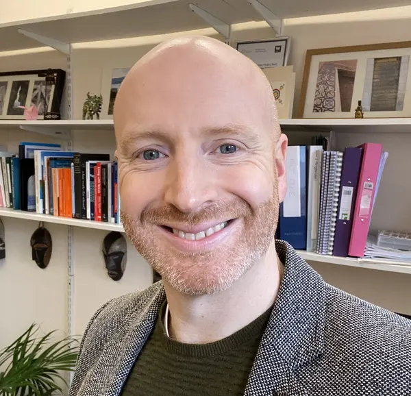 A photo of a smiling man wearing a grey blazer. He is standing in front of a bookshelf, with a green indoor plant to his right.
