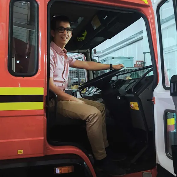 A smiling man sits in the front of a fire truck, grasping the steering wheel. The nearside door is open.