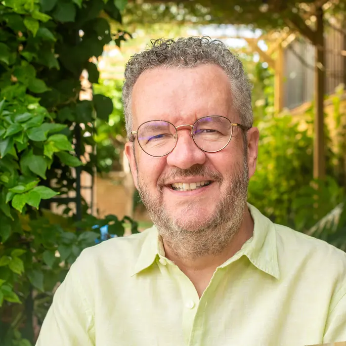 A smiling man, wearing glasses and a pale-yellow shirt, stands in front of a leafy garden.